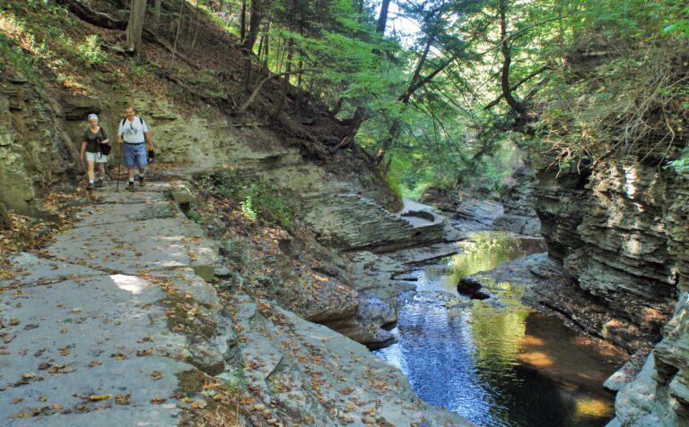 Two hikers walk on a rocky path next to a tree-lined gorge with layered stone cliffs and a shallow stream below, lit by dappled sunlight through the leaves.