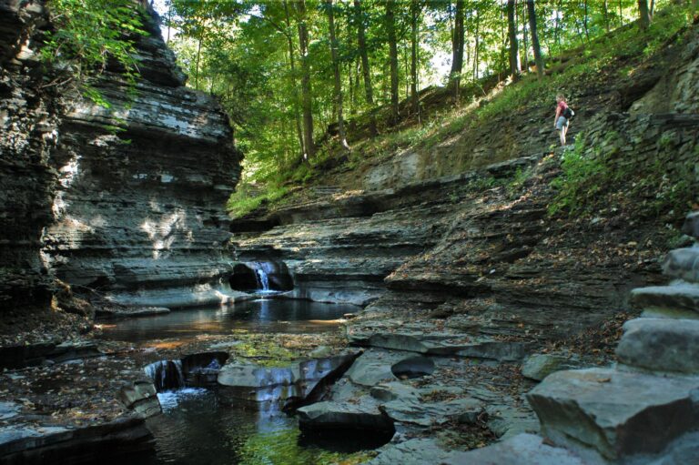 A hiker walks on a rocky trail above a gorge with layered stone walls, small waterfalls, and pools, surrounded by green forest and sunlight.