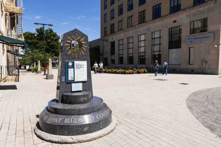 Dark stone monument with sun emblem and plaques on brick plaza near tall building; people walk by; flowers in front. Inscription partially reads: We have begun to con.