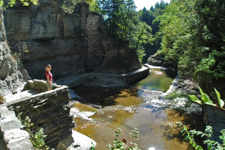 Person standing on a stone ledge above a rocky gorge with clear shallow water, surrounded by steep cliffs and green trees in bright sunlight.