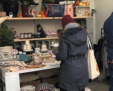 Person in a winter coat and beanie browsing a shop filled with candles, bags, gifts, and small pumpkins on decorated shelves; cozy accessories are arranged on a table in front.