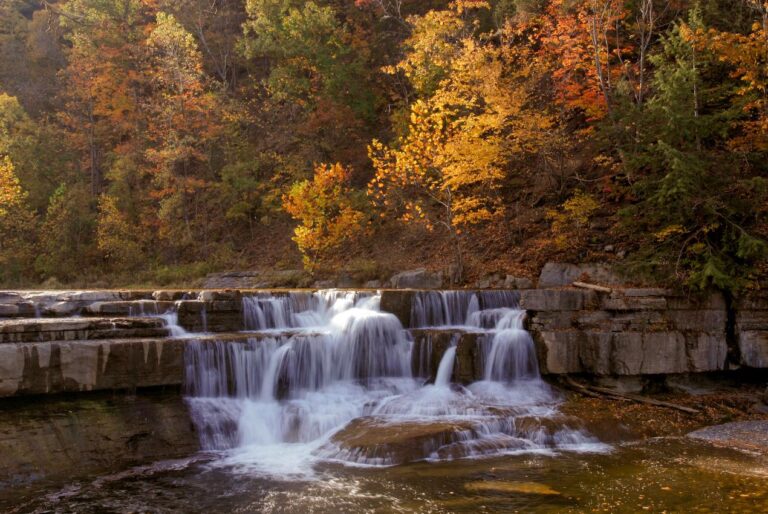 Small waterfall flowing over layered rocks, surrounded by trees with bright yellow, orange, and green autumn leaves. The water looks smooth and silky in a peaceful forest setting.