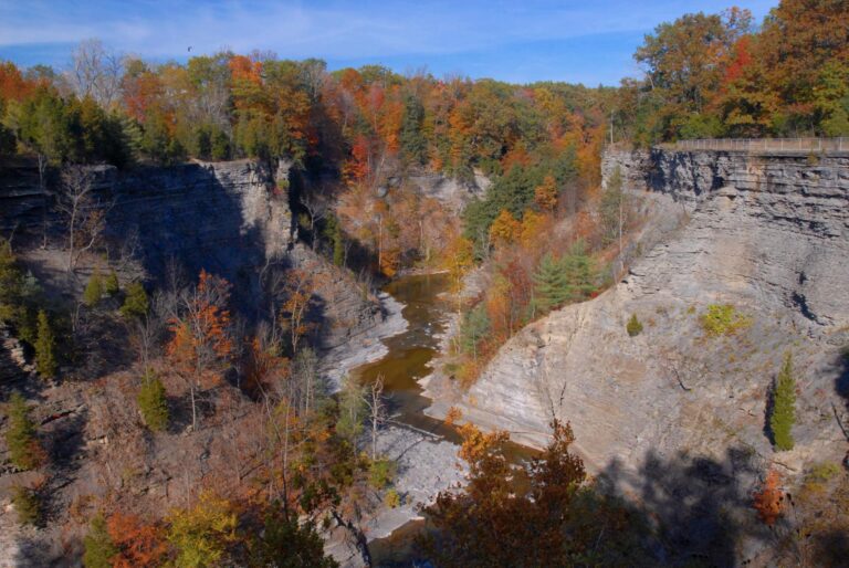 A river curves through a rocky gorge lined with dense autumn trees in red, orange, and yellow under a clear blue sky.